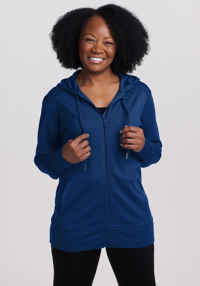 A woman with curly hair smiles in the Woolx Ryann Hoodie in Estate Blue, featuring front pockets; she pairs it with black pants against a light background—a comfy, stylish travel essential. 