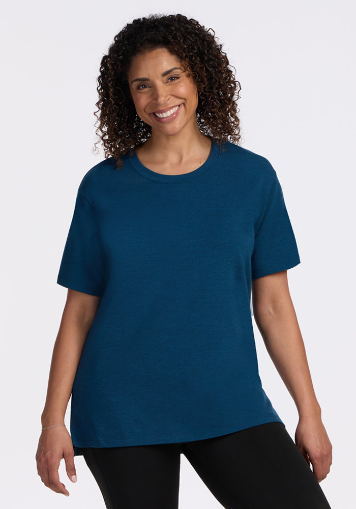 A woman with curly hair smiles while wearing the Woolx Audrey Tee in Majolica Blue and black pants, standing against a plain white background. 