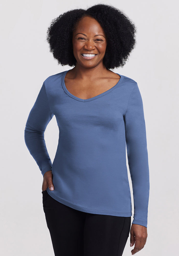 A woman with curly hair smiles while standing, wearing the Woolx Layla V Neck Top - Skyline and black pants against a plain light background. 