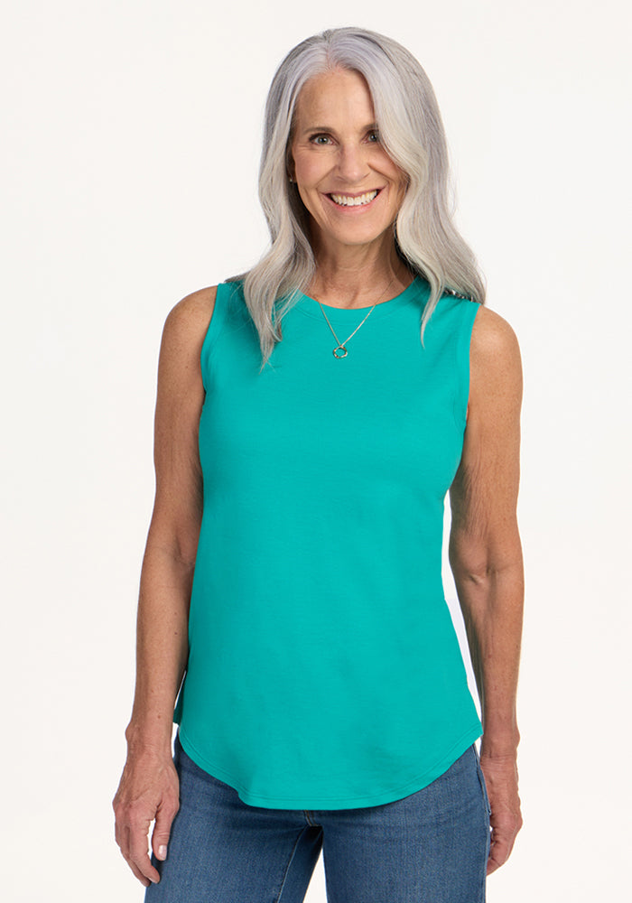 A smiling woman with long gray hair wears the Woolx Bella Tunic Top in Cool Jade and blue jeans, standing against a plain light background. She accessorizes with a necklace and rests her hands by her sides. 