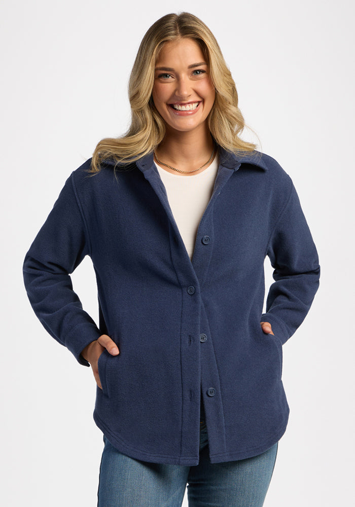 Smiling woman wears Woolx Jovie Shacket in Deep Dive, white shirt, and jeans with hands in pockets against plain backdrop. 