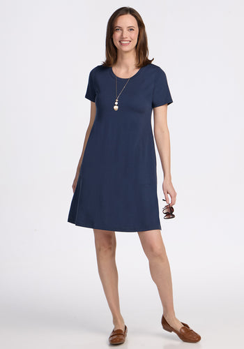 A smiling woman with straight brown hair wears the Woolx Georgie Dress in Deep Navy. She holds eyeglasses, pairs the dress with brown loafers and a long necklace, and stands against a plain white background. | Jessica is 5’10”, wearing a size S