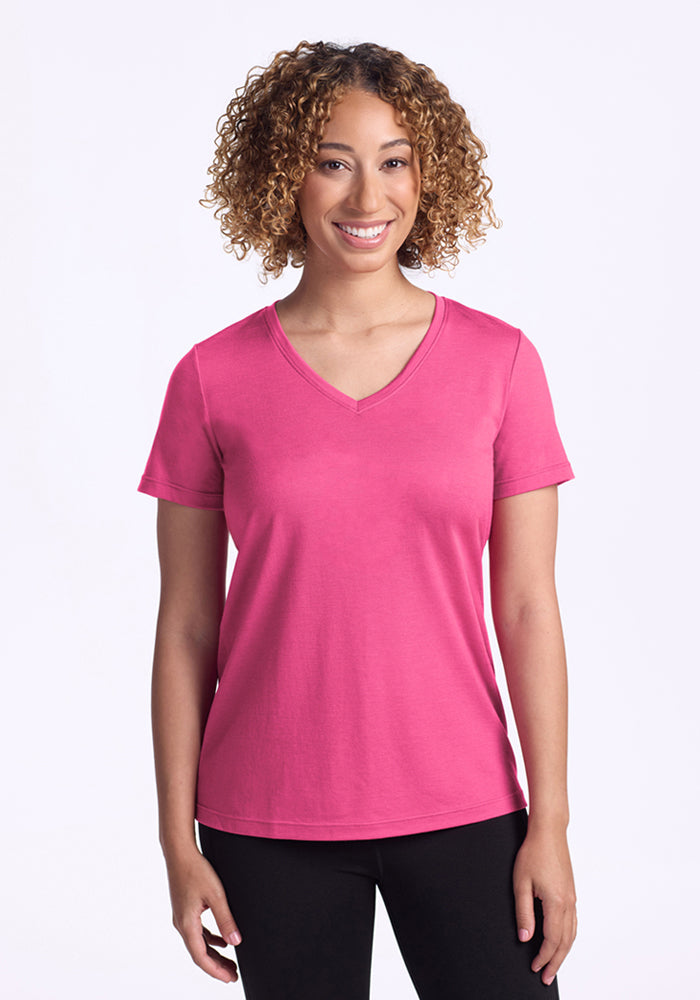 A woman with curly hair smiles while wearing the Woolx Junie V Neck in Raspberry Fizz, a relaxed fit short sleeve t-shirt, paired with black pants, standing against a plain white background. 