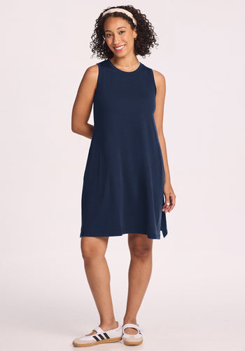 A woman with curly hair, wearing a white headband and the Woolx Clara Dress in Deep Navy, paired with white sneakers with black stripes, smiles against a plain light background. | Tori is 5'7", wearing a size S