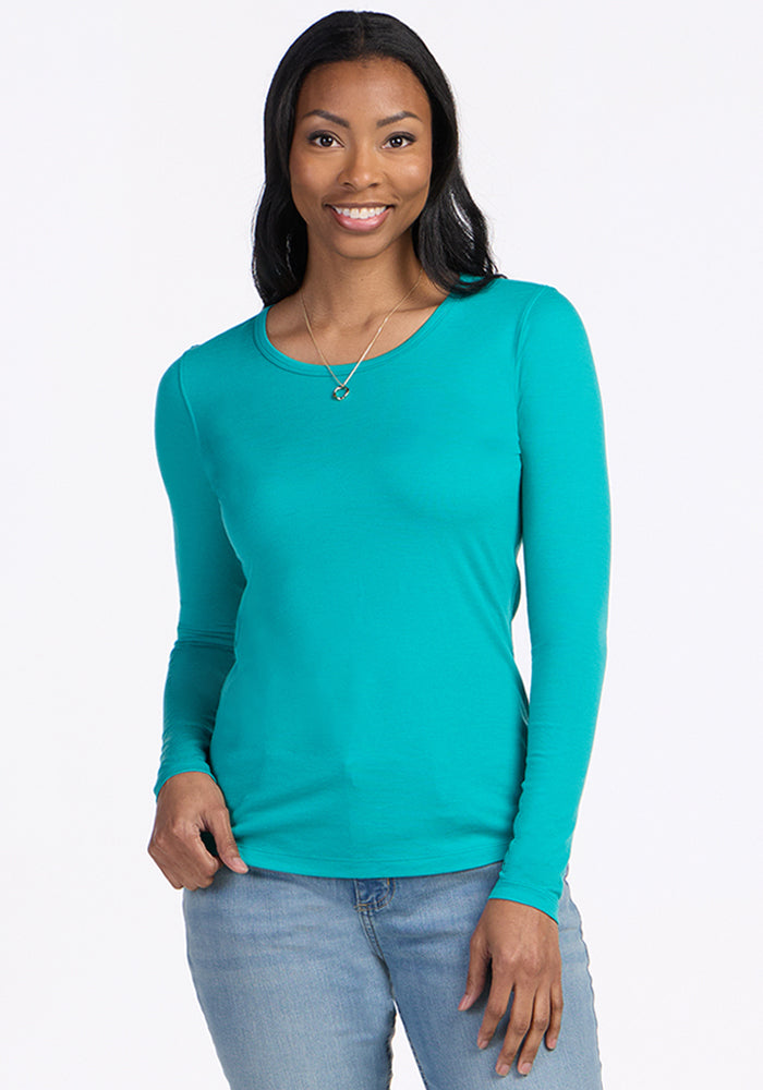 A woman with long dark hair smiles while wearing the Woolx Remi Long Sleeve T-Shirt in Cool Jade and light blue jeans. She has one hand in her pocket and stands against a plain white background. 