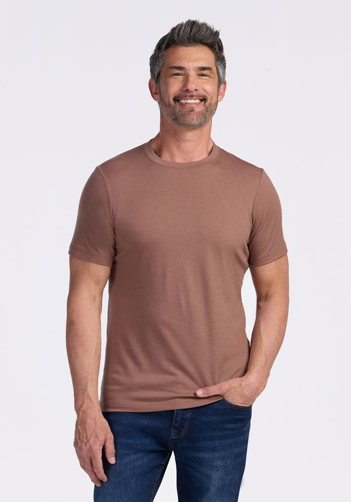 A man with short, dark hair and a beard smiles while standing in blue jeans and a Woolx Endurance Tee - Sienna Kiss, ideal performance gear, set against a plain light background. 