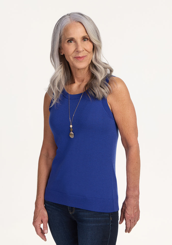 A smiling older woman with long gray hair wears the Woolx Chessy Tank Top in royal blue, paired with blue jeans and a long two-pendant necklace, standing against a plain light background. 