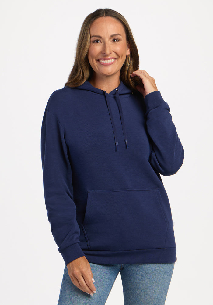 Woman in the Woolx Avery Hoodie - Ocean Cavern and jeans, smiling, touches her hair against a white background. 