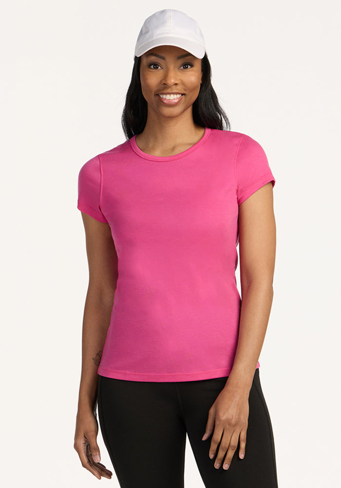 A woman wearing the Woolx Addie Short Sleeve Crew in Raspberry Fizz stands smiling, paired with black pants and a white baseball cap, against a plain white background. 