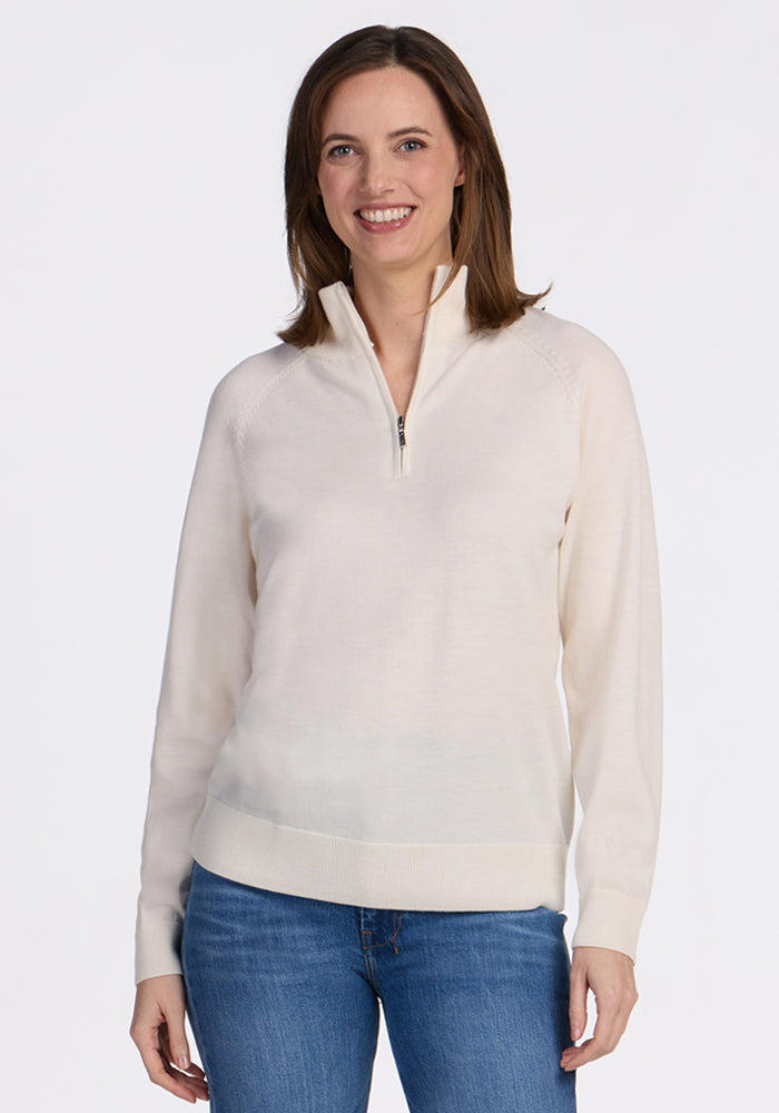 A woman with shoulder-length brown hair smiles while wearing the Woolx Collins Half Zip Sweater in Dewdrop Pearl and blue jeans, standing against a plain white background. 