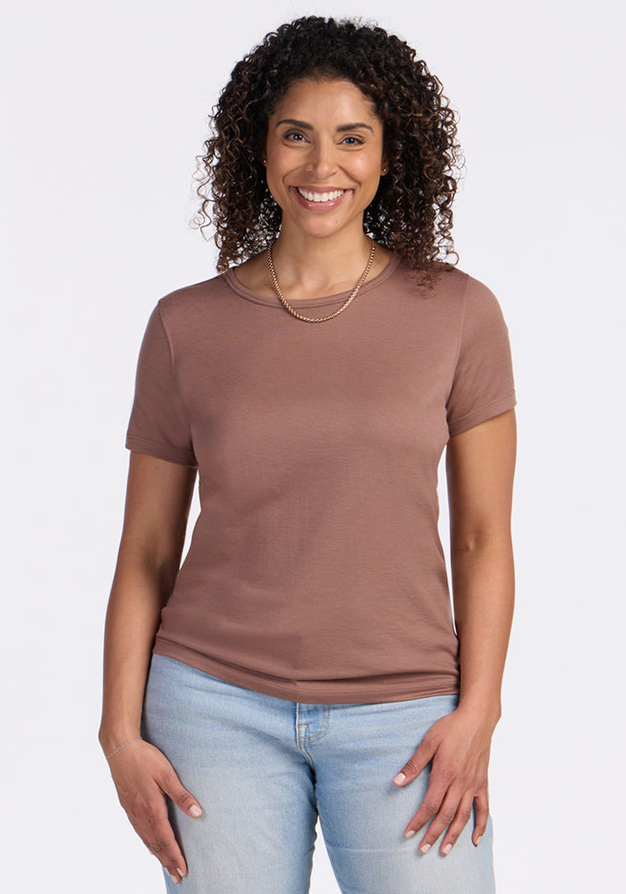 A woman with curly brown hair smiles at the camera, wearing the Woolx Liza Crew Neck Tee in Sienna Kiss, light blue jeans, and a simple gold necklace, standing against a plain white background. 