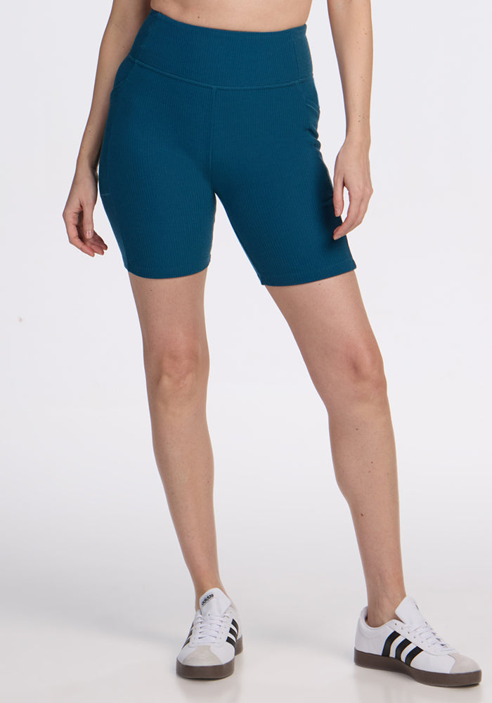 A person is shown from the waist down wearing Woolx Miley Bike Shorts in Moroccan Blue, paired with white sneakers with black stripes, standing against a plain white background. 