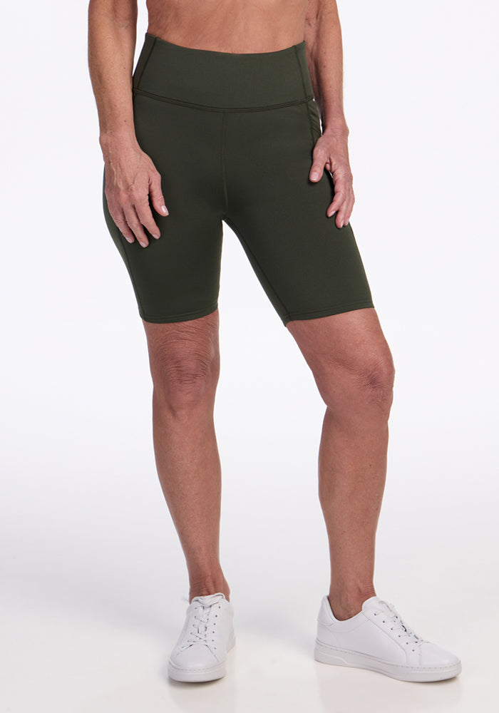 A person wearing Woolx Merino Flex® Dani Bike Shorts in Everleaf and white sneakers stands against a plain white background, with only the lower torso and legs visible. 