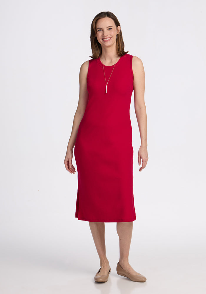 A woman smiles in the Woolx Iris Ribbed Dress in Poppy Spark, a sleeveless, knee-length bright red merino wool style, paired with a simple necklace and beige flats against a plain white background. 