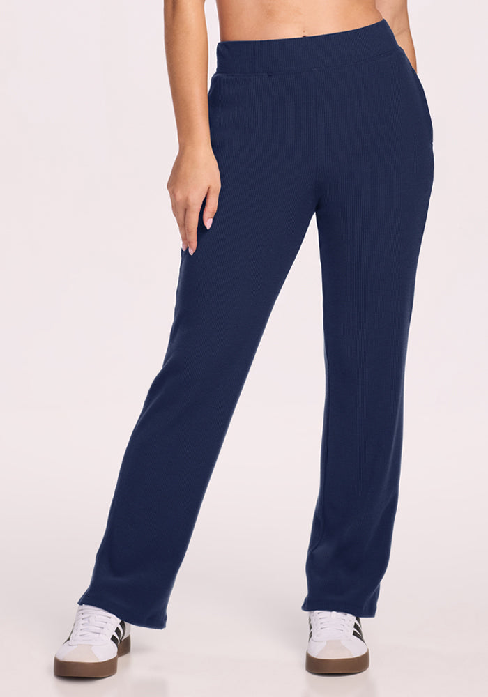 Model wears Woolx Luca Ribbed Pants in Ocean Cavern with white sneakers, hand in pocket, against a plain background. 