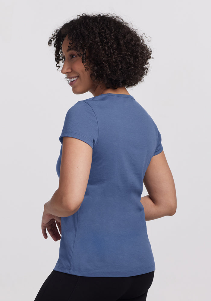 A person with short curly hair, wearing the Woolx Addie Crew Neck - Skyline and black pants, smiles and looks over their shoulder against a plain light background.