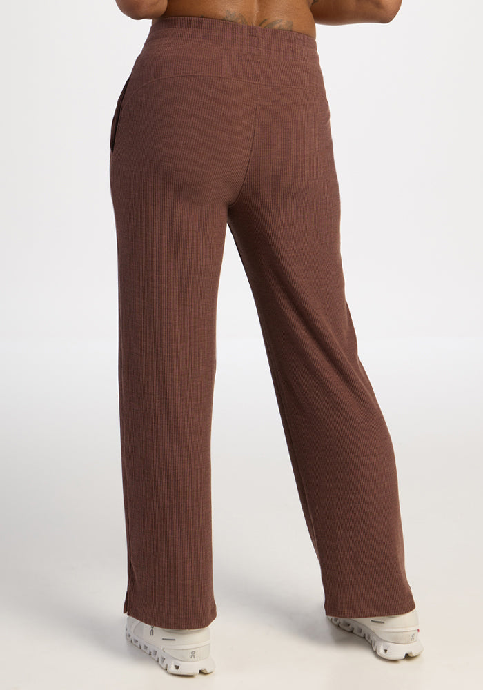 Model wears Woolx Luca Ribbed Pants - Sable Heather with white sneakers, seen from behind on a plain white background.