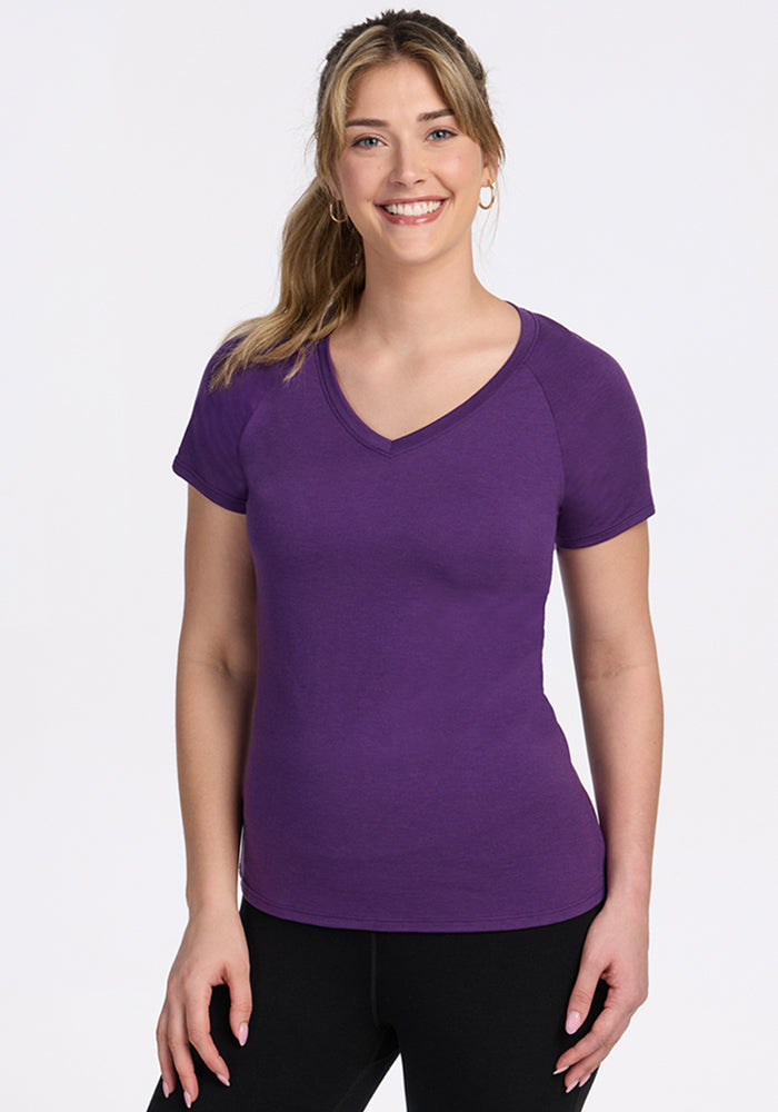 A woman with long, light brown hair in a ponytail smiles against a white background, wearing the Woolx Mia V Neck t-shirt in Plum Crush and black pants. 