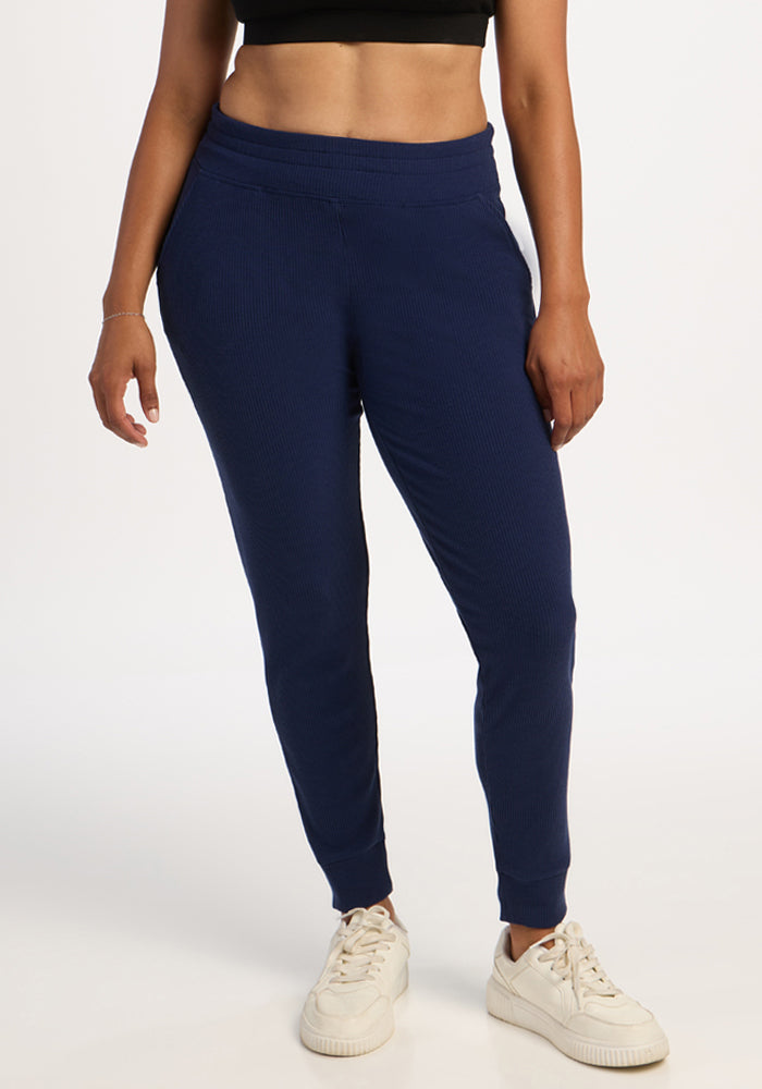 Woman wears Woolx Lennon Ribbed Joggers in Ocean Cavern, black crop top, and white sneakers against plain background. 