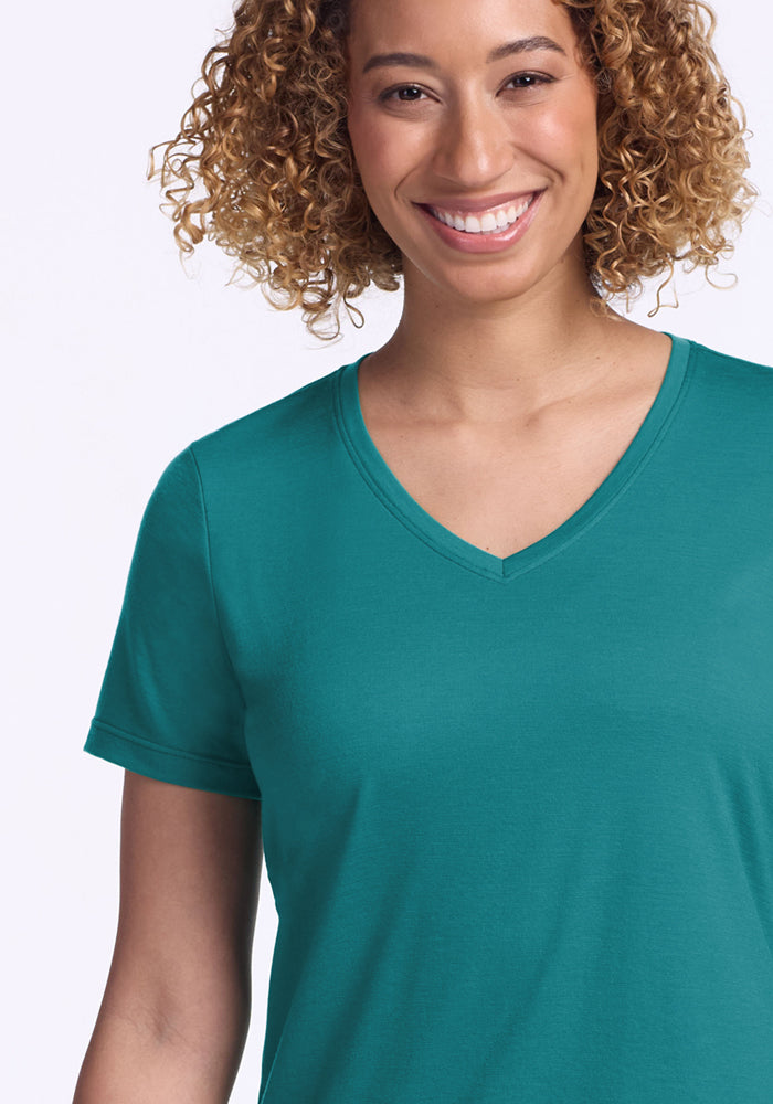 A curly-haired woman smiles in the Woolx Junie V Neck - Surfside, a relaxed-fit teal Merino wool tee for women that resists odor, against a plain light background.