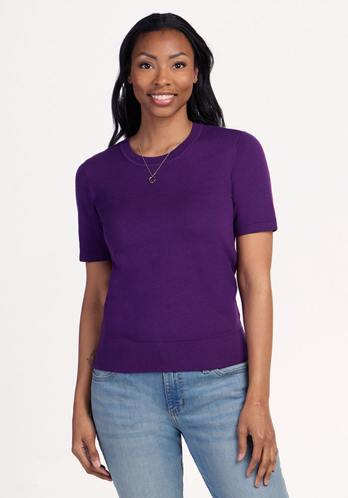A woman with long dark hair smiles at the camera, wearing the Woolx Anna Crew Sweater - Plum Crush paired with light blue jeans and a gold necklace, standing in a relaxed pose against a plain light background. 