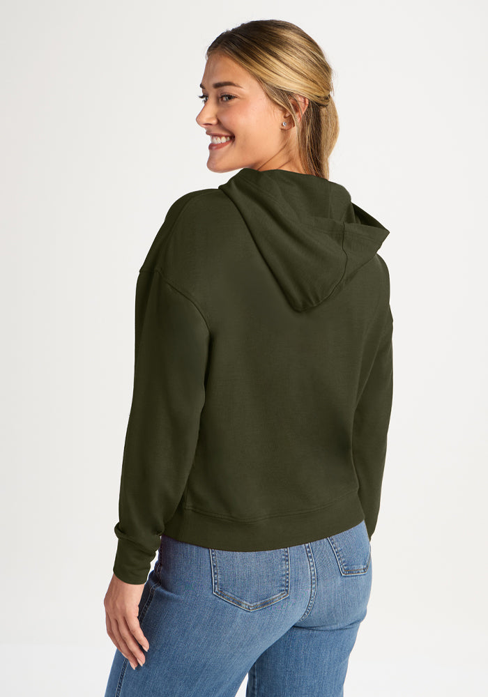 Woman smiles, looking back in a Woolx Lizzy Hoodie - Everleaf and blue jeans against a plain light background.