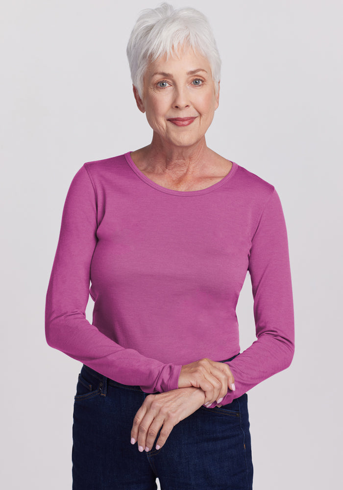 Smiling older woman with short white hair wears the Woolx Remi Long Sleeve T-Shirt in Purple Wine and dark jeans, standing against a light gray background. This merino top is moisture-wicking and odor-resistant. 