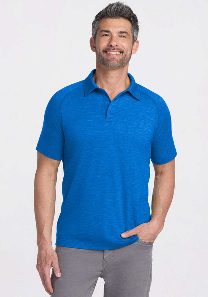 A man with short gray hair and a beard is smiling, wearing the Woolx Summit Polo in Glacial Blue and light gray pants. He poses with one hand in his pocket against a plain background, highlighting the polo’s moisture management. 