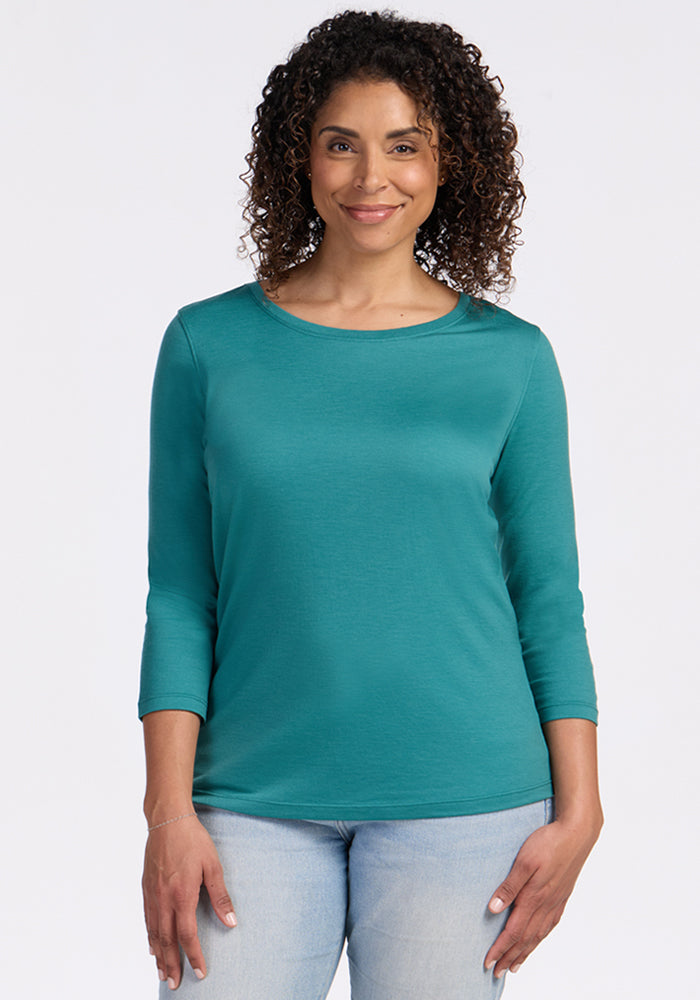 A woman with curly hair wears the Woolx Jenny 3/4 Sleeve Crew Neck in Surfside and light blue jeans, standing forward and smiling against a plain white background. 