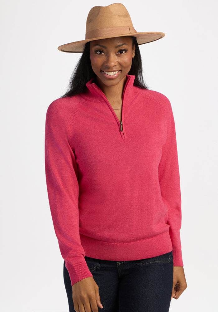 Woman in a tan wide-brim hat, Woolx Collins Half Zip Sweater in Vivid Rose, and jeans smiles by a white backdrop. 