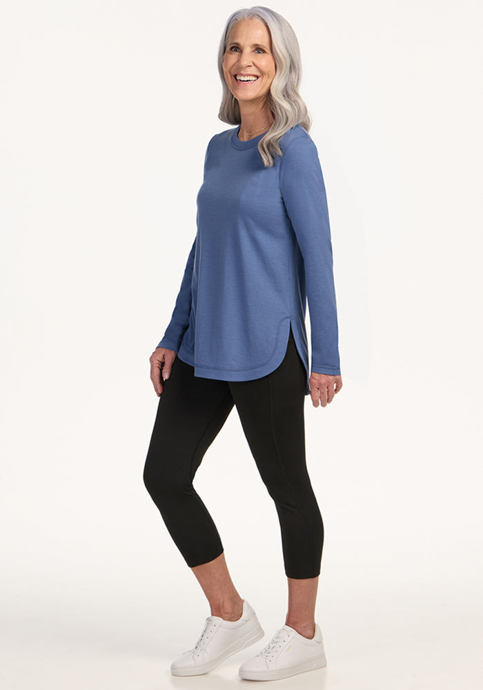 A smiling older woman with long gray hair wears the Woolx Hazel Tunic - Skyline, featuring lightweight Merino wool, black cropped leggings, and white sneakers as she stands against a plain light background.