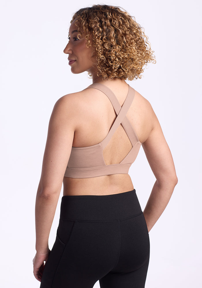 A woman with curly hair is shown from the back, wearing the Woolx Livi Bra in Morning Latte and high-waisted black leggings, standing against a plain white background.