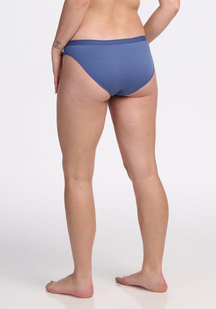 A person stands barefoot on a white surface, seen from the back, wearing blue Woolx Roxie Bikini Underwear - Skyline and showing a small tattoo on their left arm.