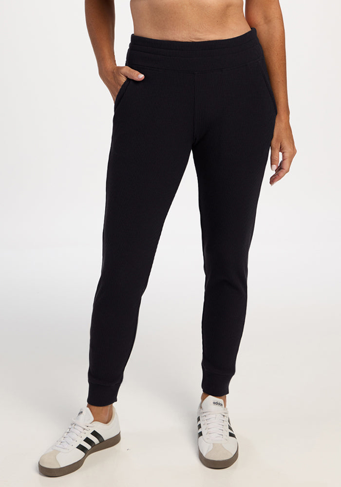 Model in Woolx Lennon Ribbed Joggers - Black and white sneakers, hands in pockets, on a white background. 
