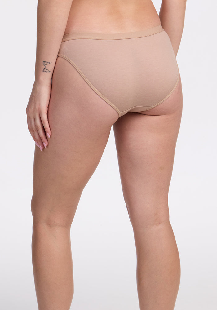 A person stands with their back to the camera, wearing Woolx Roxie Bikini Underwear in Morning Latte. Their left hand is by their side, showing a small wrist tattoo, against a plain light-colored background.