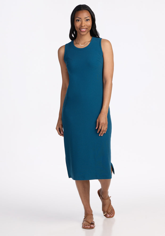 A woman stands against a plain white background, wearing the Woolx Iris Ribbed Dress in Moroccan Blue with brown sandals. She smiles softly, one arm by her side and the other hand resting on her hip.