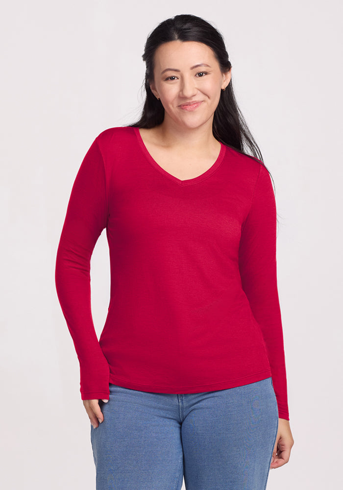 A woman with long dark hair wears the Woolx Millie Long Sleeve in Poppy Spark and blue jeans, standing against a plain light background and smiling gently at the camera.