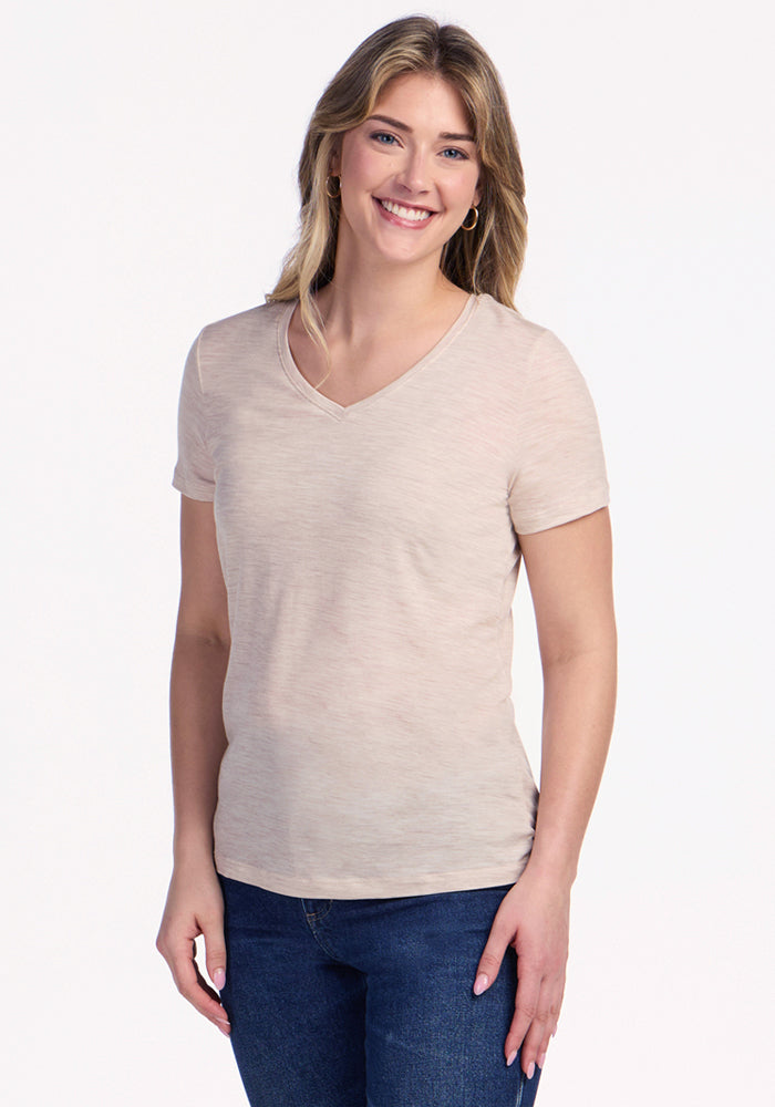 A smiling woman with long, light brown hair wears the Woolx Junie V Neck in Cream Heather and blue jeans, standing against a plain white background. 