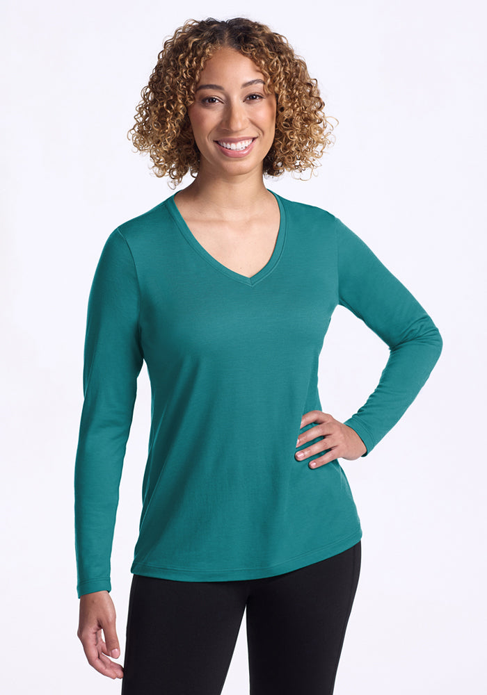 A smiling woman with curly hair wears the Woolx Millie Long Sleeve - Surfside and black pants, standing confidently against a plain white background with one hand on her hip. 