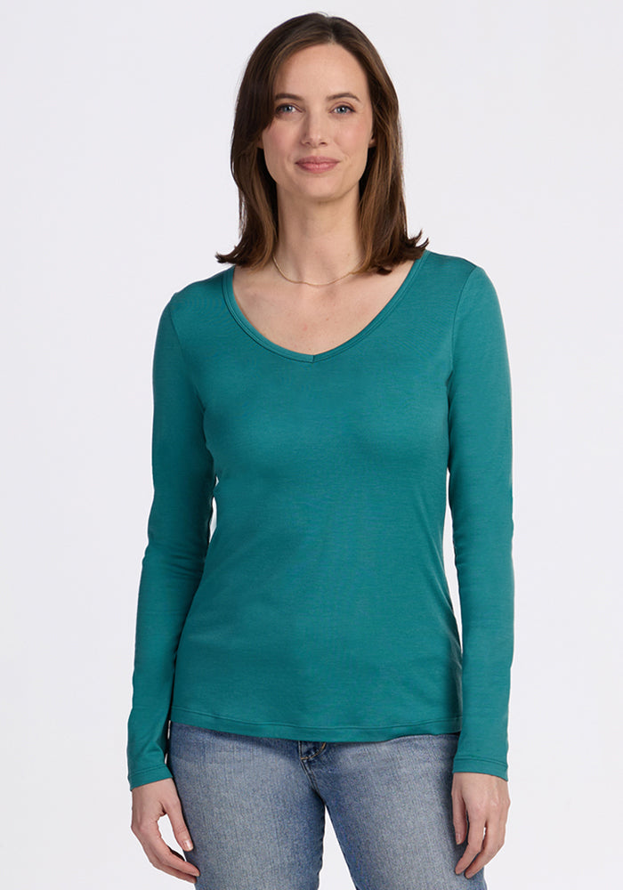 Woman with straight brown hair wears the Woolx Layla V Neck Top in Surfside with blue jeans, smiling at the camera. This odor-resistant, moisture-wicking merino wool shirt brings comfort and style against a plain white background. 