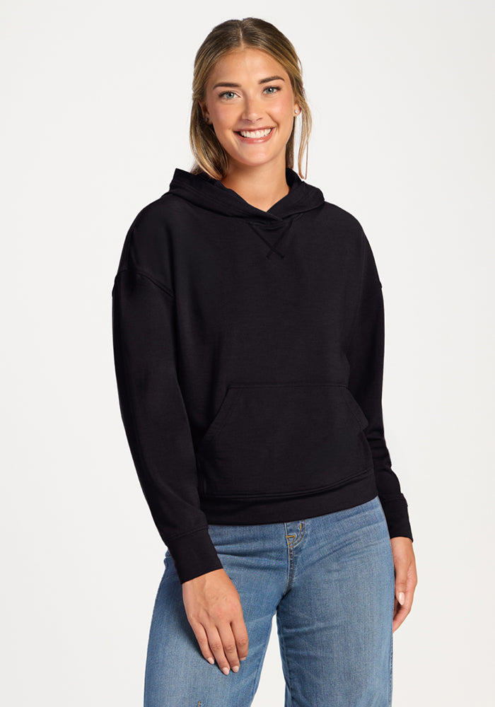 Smiling woman wears the Woolx Lizzy Hoodie in black with blue jeans, standing against a plain white background.