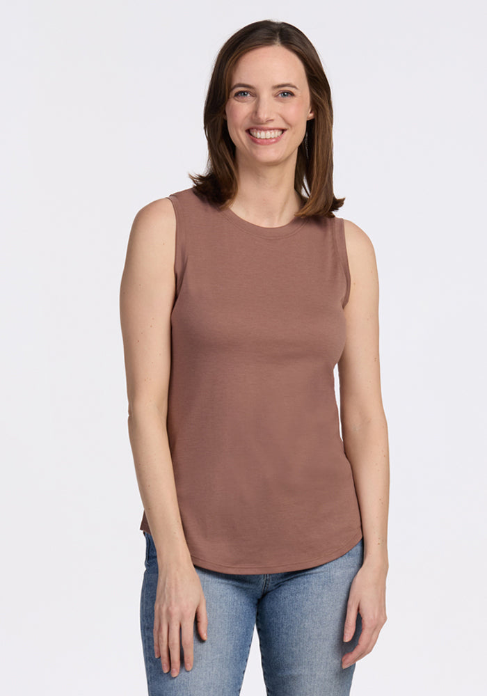 A woman with shoulder-length brown hair smiles while standing against a plain white background, wearing the Woolx Bella Tunic Top in Sienna Kiss and blue jeans. 