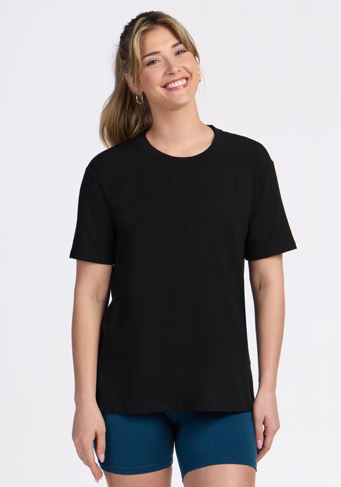 A woman with long, light brown hair smiles in a relaxed fit Woolx Audrey Tee - Black and blue shorts, standing against a simple white background. 