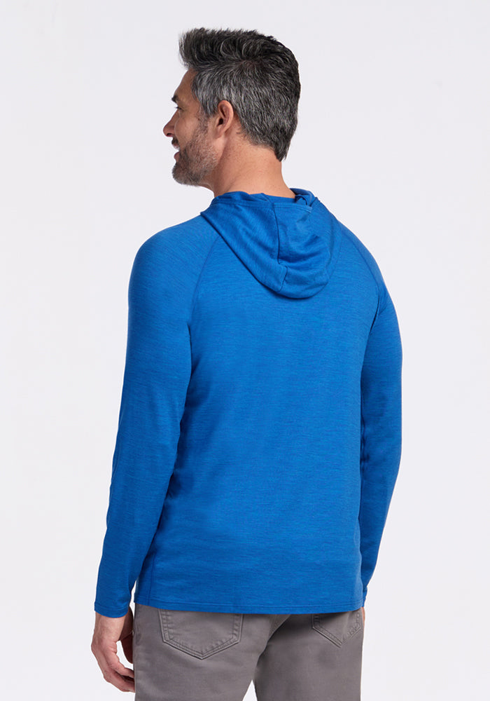 A man with short dark hair and a beard, seen from behind, wears the Woolx Base Camp Hoodie in Glacial Blue and gray pants, highlighting comfortable, lightweight outdoor apparel against a plain white background.