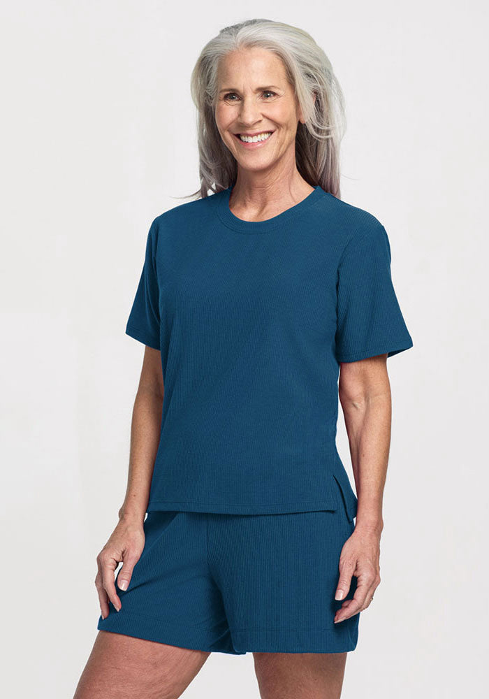 A smiling older woman with long gray hair wears the Woolx Delia Ribbed Short Sleeve in Moroccan Blue and matching blue shorts, standing against a plain white background. 