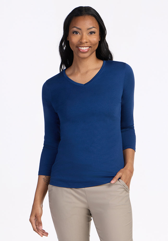 A woman with long dark hair smiles while wearing the Woolx Elena 3/4 Sleeve V Neck in Estate Blue and beige pants, standing against a plain white background.