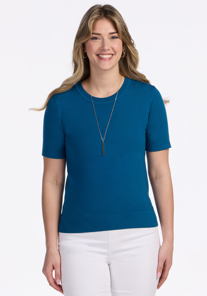 A woman with long, wavy blonde hair smiles while wearing the Woolx Anna Crew Sweater in Moroccan Blue, paired with white pants and a pendant necklace, standing against a plain white background. 