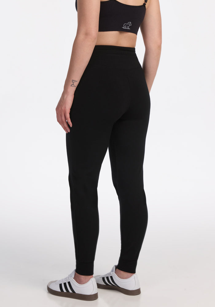 A person in Woolx Madden Tall Joggers - Black, a black sports bra, and white sneakers stands against a white backdrop.