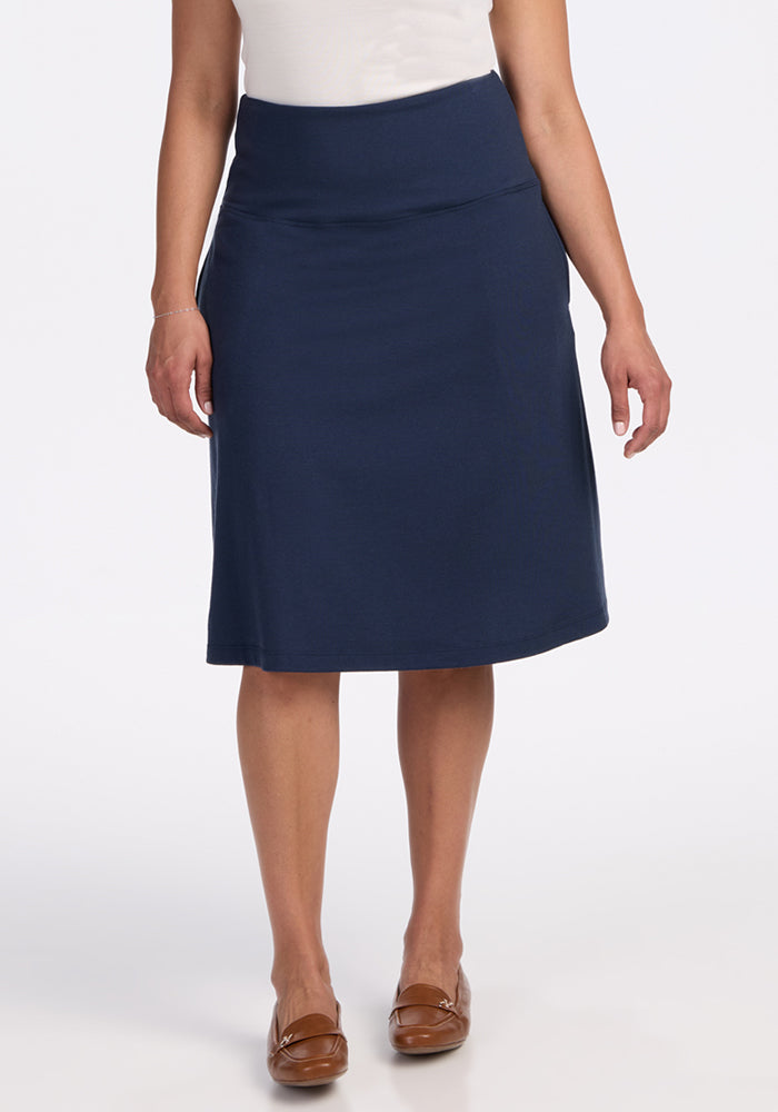 A person stands facing forward in a white top and the Woolx Marilyn Flowy Midi Skirt in Deep Navy, paired with brown flats. The background is plain white. 