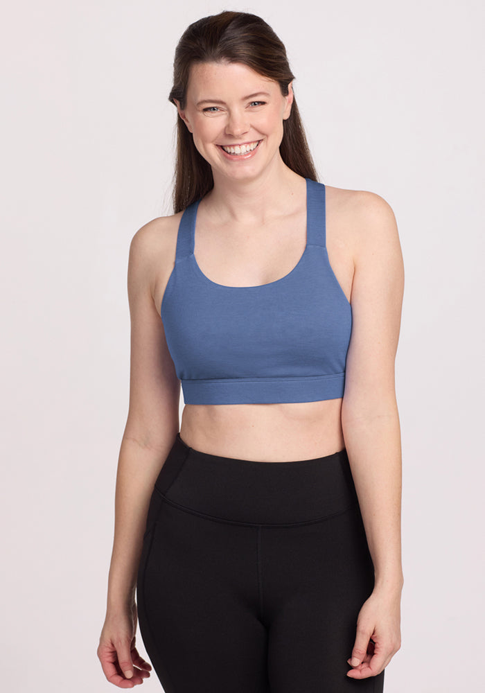 A woman with long brown hair, wearing the Woolx Livi Bra - Skyline and black leggings, stands smiling against a plain light background. 